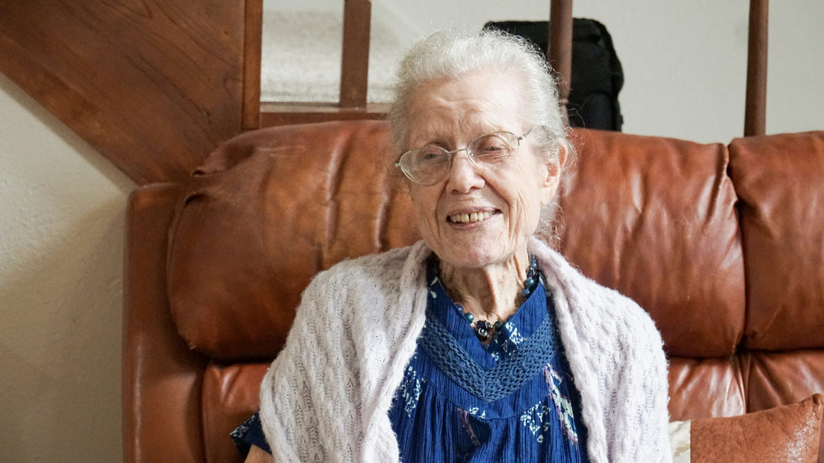 Femme âgée souriante portant des lunettes, un cardigan clair et un collier bleu