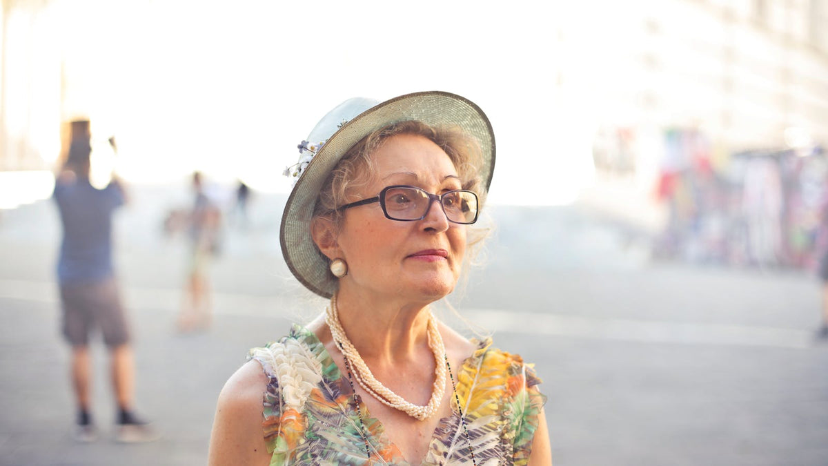 Femme de plus de 60 ans portant lunettes, rouge à lèvres rose, chapeau et collier perlé Bild